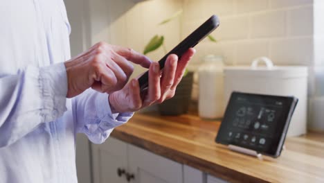 sección media de una mujer caucásica mayor de pie en la cocina, usando un teléfono inteligente y una tableta