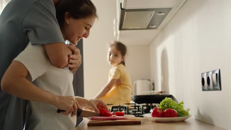 A-man-in-a-gray-T-shirt-approaches-his-brunette-girl-wife-and-hugs-her-while-preparing-morning-breakfast-and-salad-near-his-daughter,-brunette-girl-in-a-yellow-dress-in-the-kitchen-in-the-morning