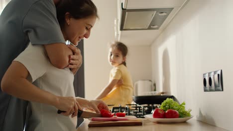un hombre con una camiseta gris se acerca a su esposa morena y la abraza mientras prepara el desayuno y la ensalada de la mañana cerca de su hija, una chica morena con un vestido amarillo en la cocina por la mañana