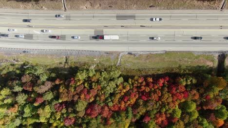 Top-down-aerial-overhead-view-of-busy-interstate-highway-that-cuts-through-forest-during-autumn,-leaves-display-peak-fall-foliage