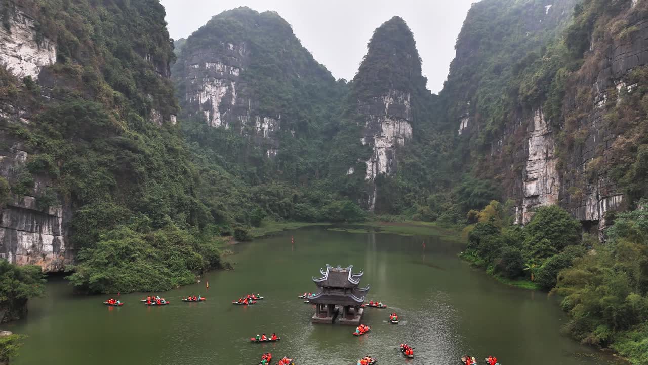 Tourists paddle boats along the green river at Trang An, Ninh Binh, Vietnam, encircling a small temple set against dramatic limestone karst mountains covered in lush vegetation under a cloudy sky
