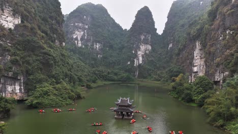 tourists paddle boats along the green river at trang an, ninh binh, vietnam, encircling a small temple set against dramatic limestone karst mountains covered in lush vegetation under a cloudy sky