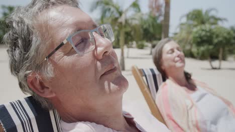 happy senior caucasian couple sitting in deckchairs asleep on sunny beach, in slow motion