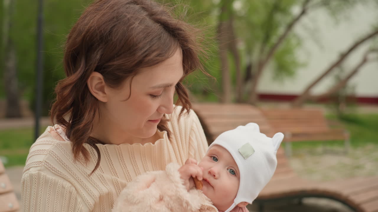 White Mother Feeding Baby Cookie On Park Bench With Cozy Pink Outfit And Curious Infant Reaching For Snack Tender Playful Exchange, Food Reaction, Outdoor Trees And Affectionate Interaction