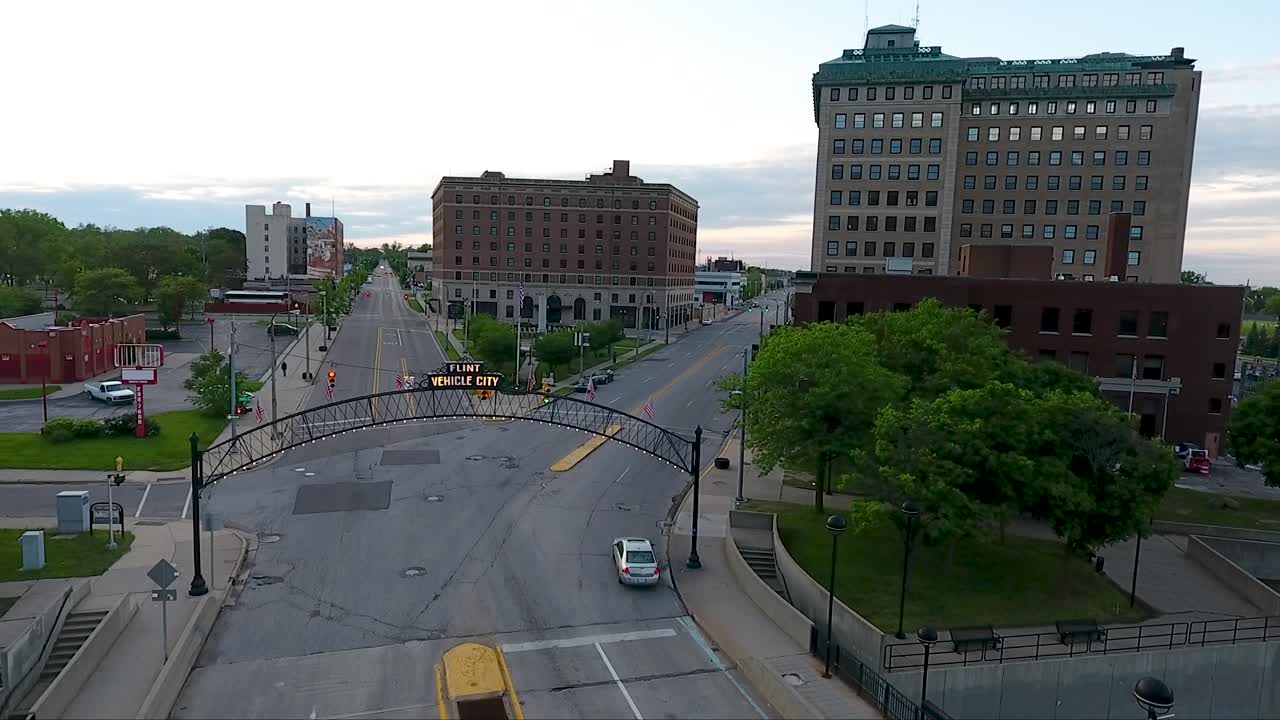 A drone flies in reverse, showing the old brick buildings of downtown Flint, Michigan and the lighted Vehicle City Sign and archway above South Saginaw Street and revealing the Flint River.