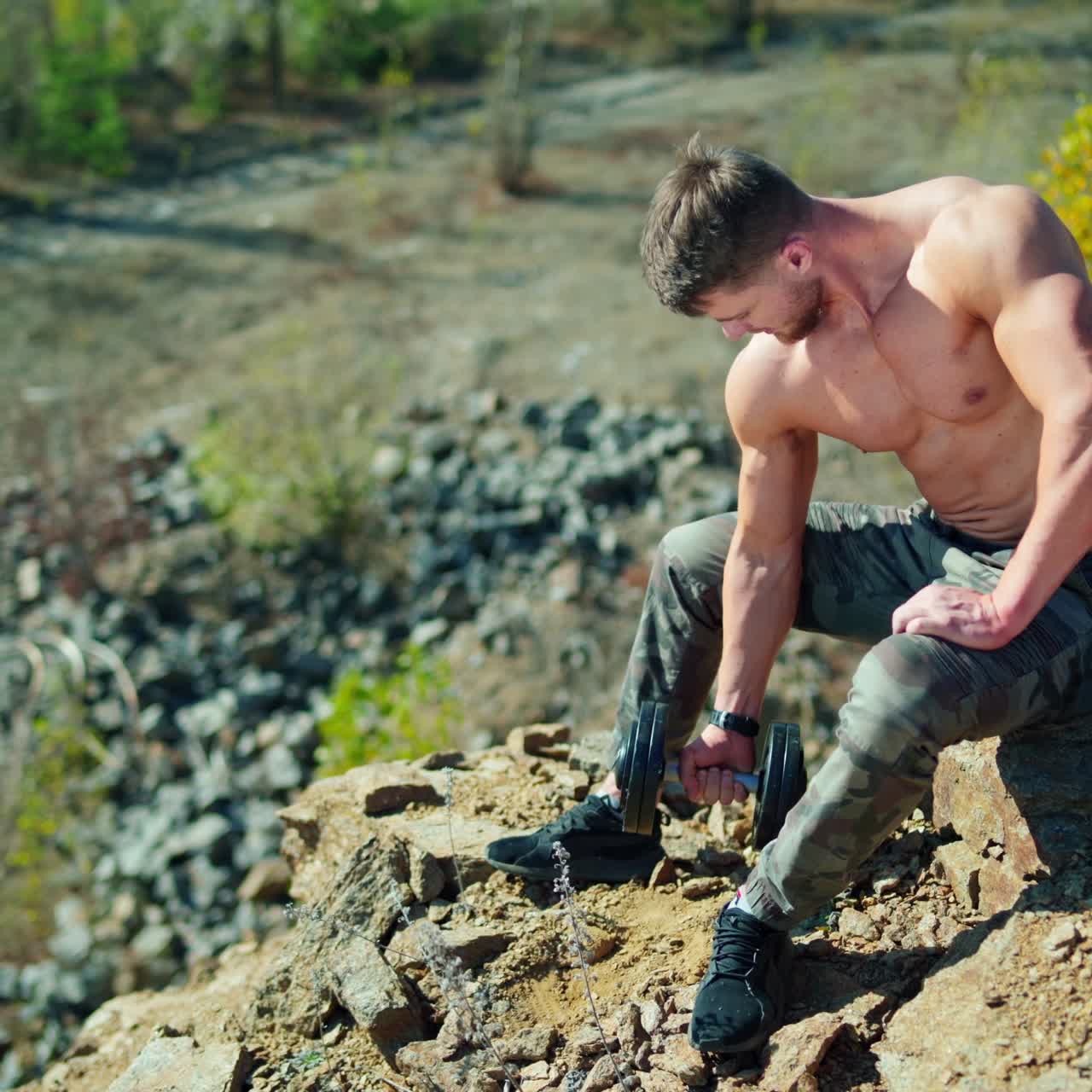 Handsome sportsman with a dumbbell outdoors. Muscular athlete pumping his arm biceps on the rocky background in summer day.