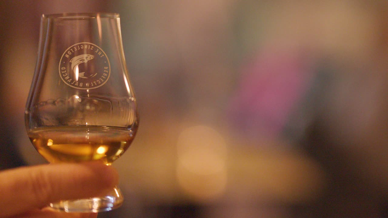 A hand gently swirls a whisky glass, highlighting golden liquid and reflections in a warmly lit, softly blurred pub or bar environment