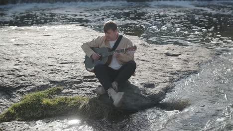 young man playing guitar sitting on the bank of a mountain river on a background of rocks. concept of freedom relaxation. place