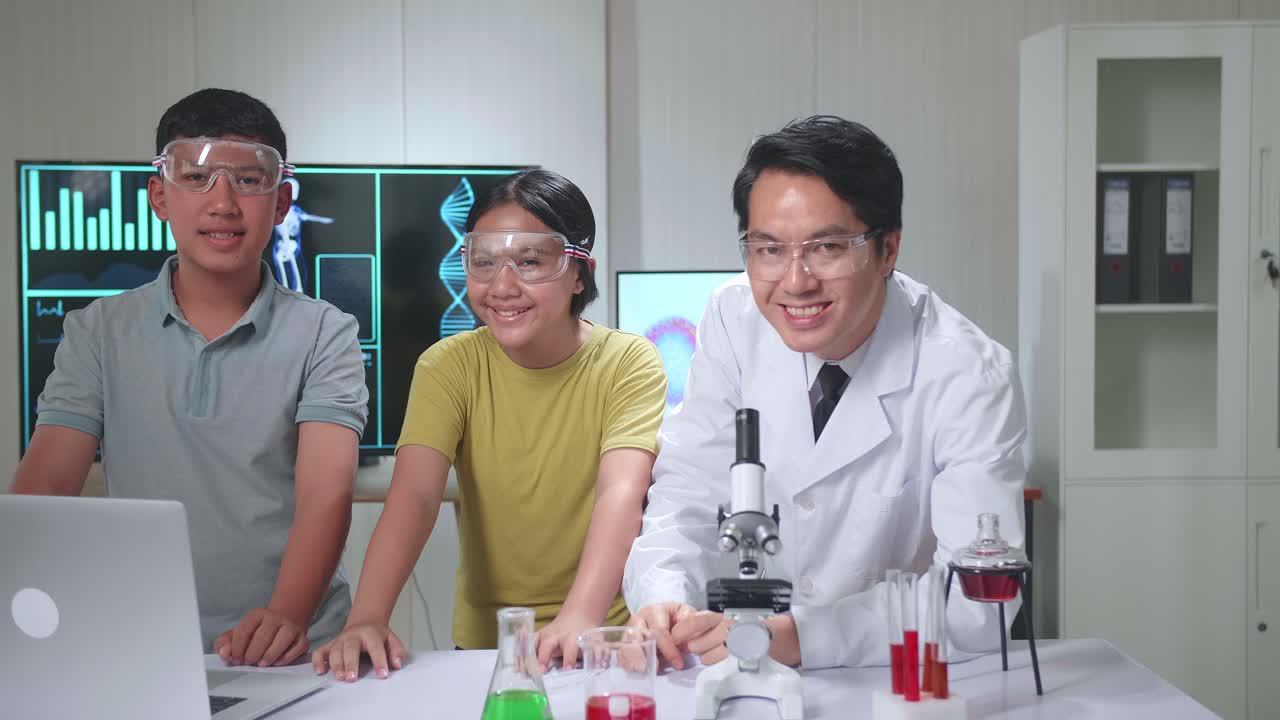 Young Asian Boy And Girl Learning Science Experiment In Laboratory With Teacher In Classroom, They Smile To Camera