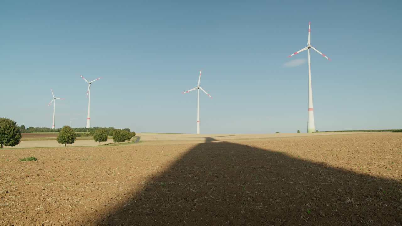 Four wind turbines stand tall on a sunny day, their shadows stretching across a freshly plowed agricultural field under a clear blue sky