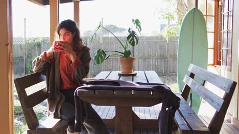 mujer de raza mixta pensativa sentada a la mesa bebiendo té en la cocina de una cabaña soleada