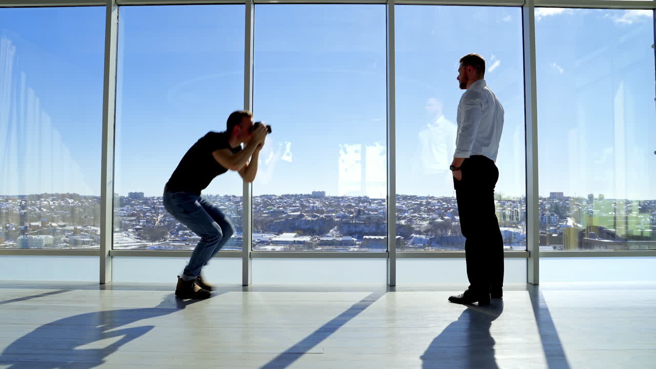 Photographer taking photos of a male model. Young businessman posing on camera in office near the large windows on city background. Side view.