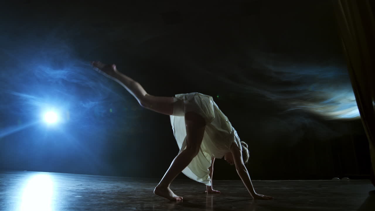 baila moderna mujer en un vestido blanco baila un ballet moderno salta en el escenario con humo en los reflectores azules.