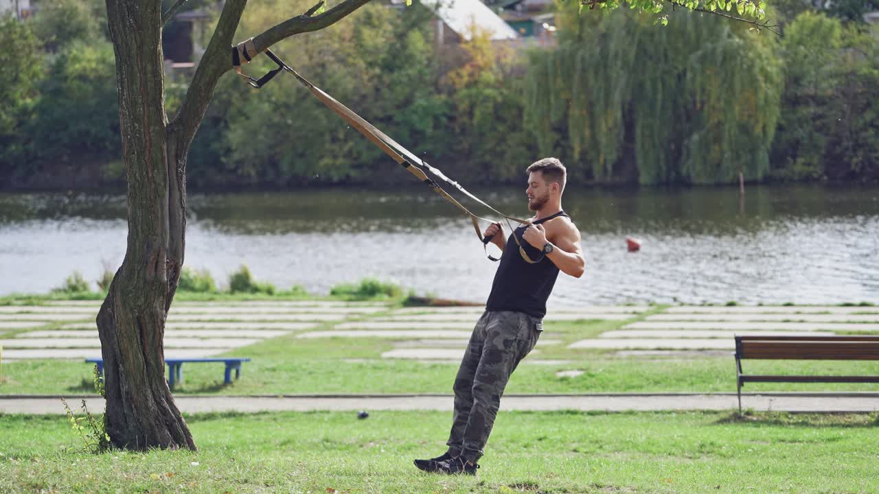 Man doing workout on trx near the river in summer. Young male exercising outdoors with straps. Side view of an athlete doing bungee training.