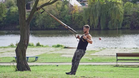 man doing workout on trx near the river in summer. young male exercising outdoors with straps. side view of an athlete doing bungee training.