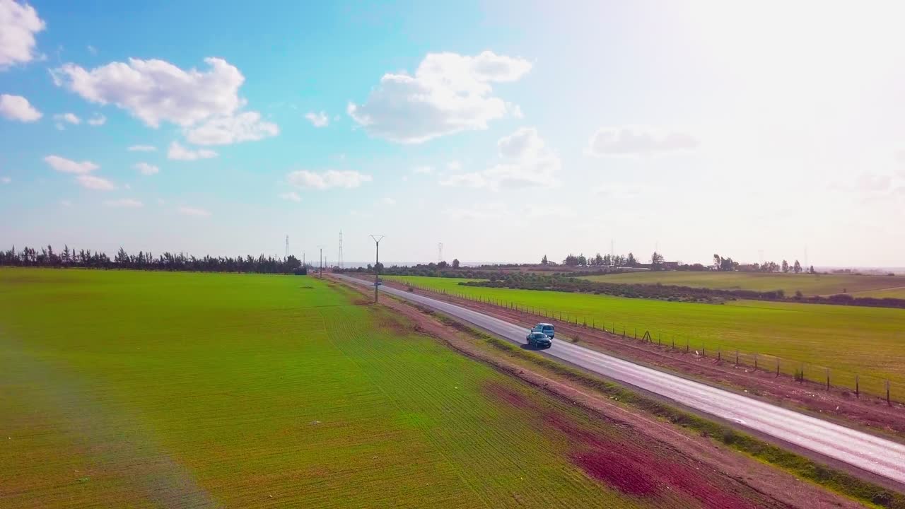A beautiful aerial shot shows a single car driving down a remote road, bordered by vast green fields under a bright blue sky