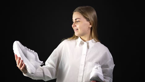 lady in white shirt holds white sneakers from different pairs in both hands. woman is making choice between similar shoes. black backdrop.