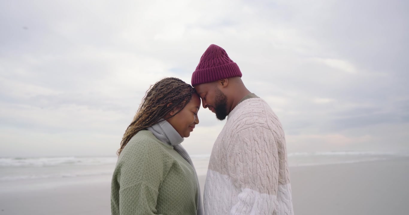 Romantic couple holding hands on shore by ocean, man in beanie closing distance, pressing foreheads