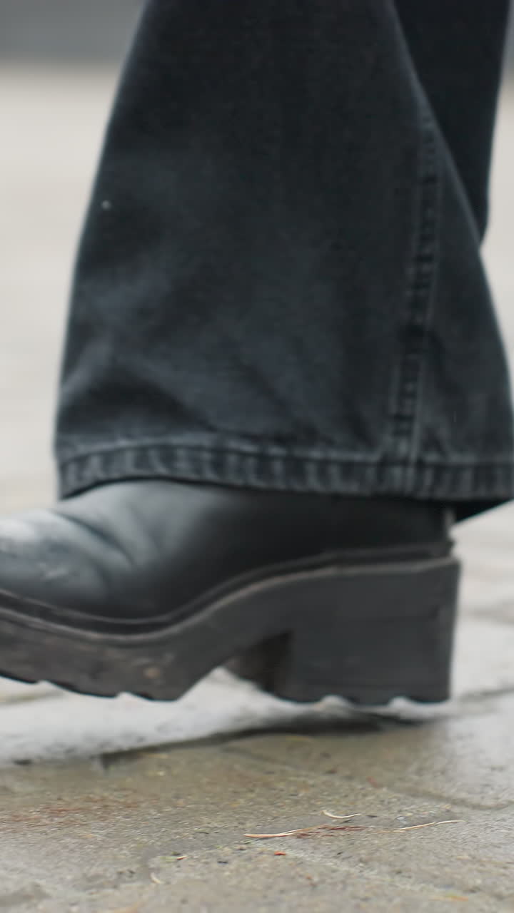 Close up of person's leg wearing black trousers and black boots walking slowly forward on outdoor paved path during calm overcast day, emphasizing footwear motion and moody atmosphere
