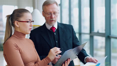 young secretary talking to a senior businessman. beautiful woman in glasses is showing new ideas on a paper to her boss on the office background.