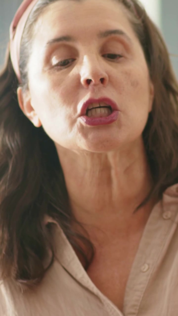 Video verticale: Uomo con maglietta rossa che si appoggia e ascolta, mentre una donna con camicetta color cuoio entra e discute a casa, con una pianta.