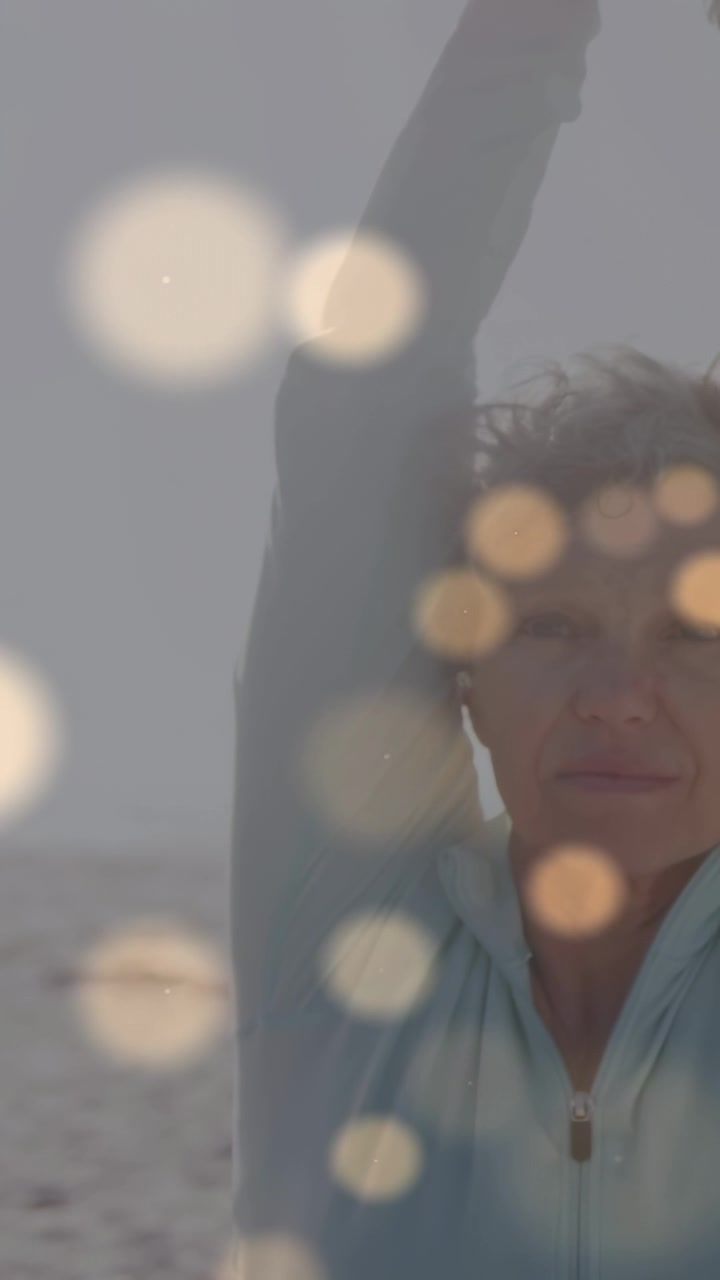 Video verticale: Donna matura che alza il braccio e pratica yoga sulla spiaggia per la calma, il sole crea un effetto bokeh.