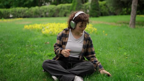 Happy-brunette-girl-in-a-brown-shirt-sits-on-the-lawn-and-turns-on-music-on-wireless-headphones-in-the-park