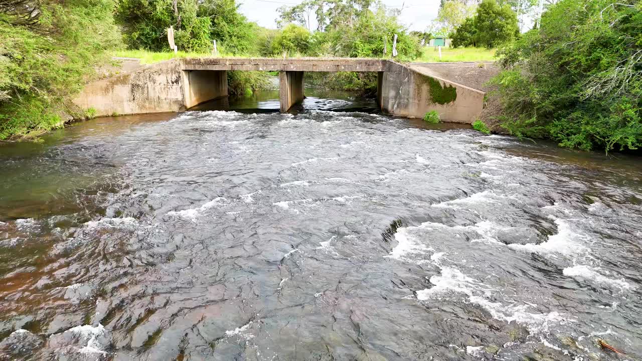 Camera slowly pans over fast-moving stream under bridge, lush greenery, daylight, steady movement