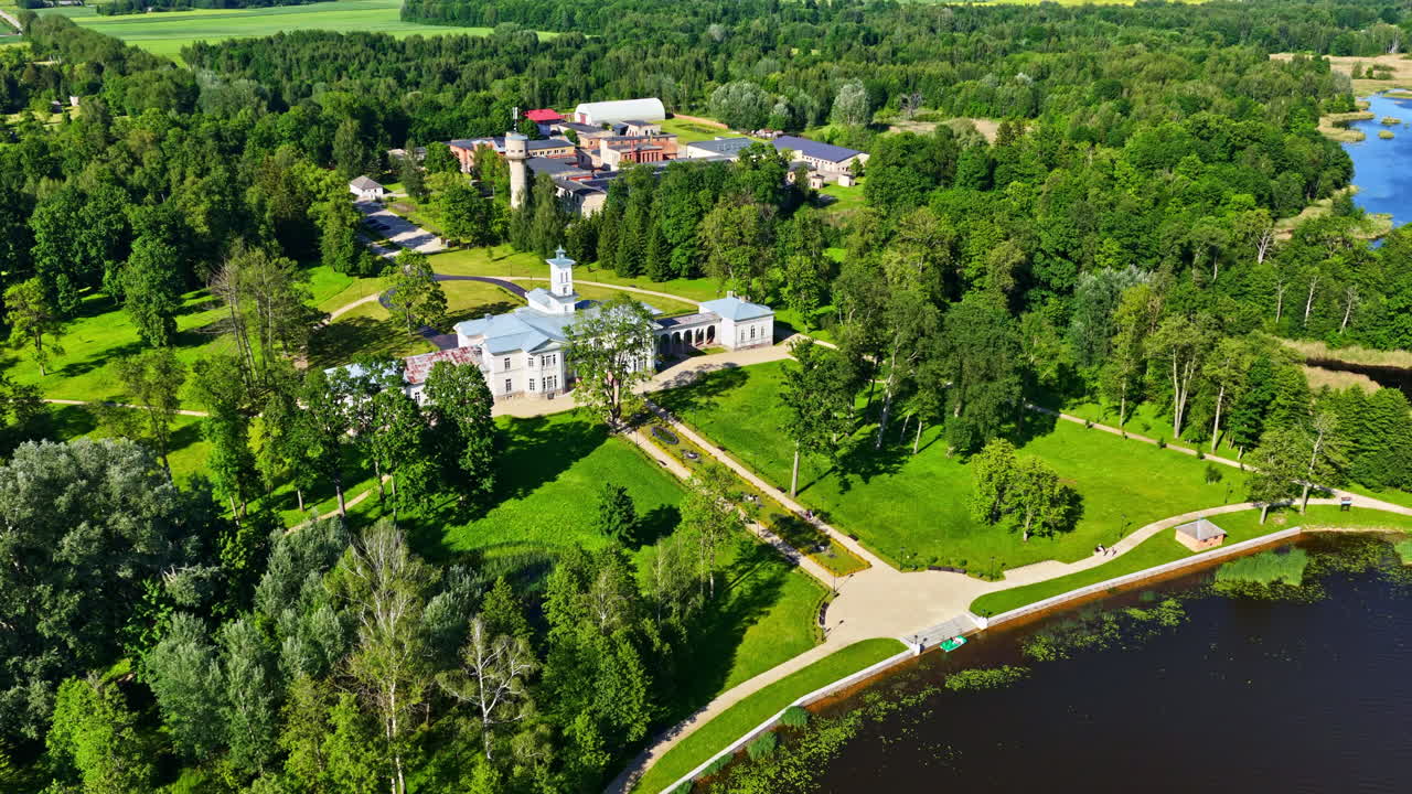 Aerial parallax movement reveals Astravas Manor, an Italian villa-style palace, nestled in lush parkland beside a scenic lake in Biržai, Lithuania