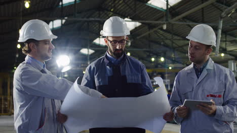 trois ingénieurs portant des casques tenant un plan et une tablette tout en parlant dans une usine