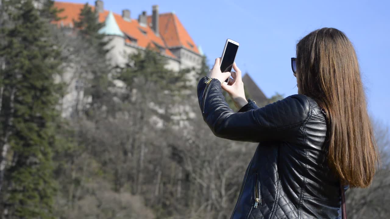 una encantadora joven con chaqueta de cuero negra tomando fotografías de un castillo en su teléfono en un parque público en un agradable día soleado de primavera