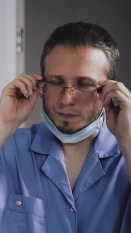 portrait of young shaggy unshaven doctor, dressed in surgical care suit, looks puzzled in mirror at his reflection, takes off his glasses, protective mask, anxiously ponders exit from quarantine