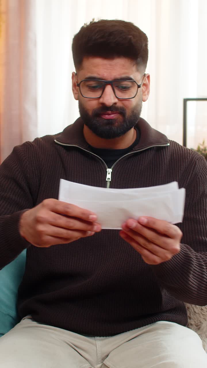 Indian man looking through receipts with worry on sofa at home financial stress and anxiety visible