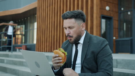 businessman eating sandwich on stairs while working on laptop