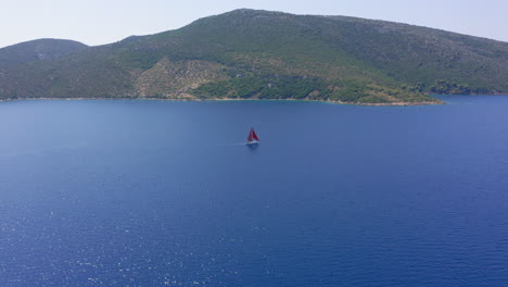 aerial: slow orbit drone shot of a sailboat with red sails that is sailing offshore on a clear summer day