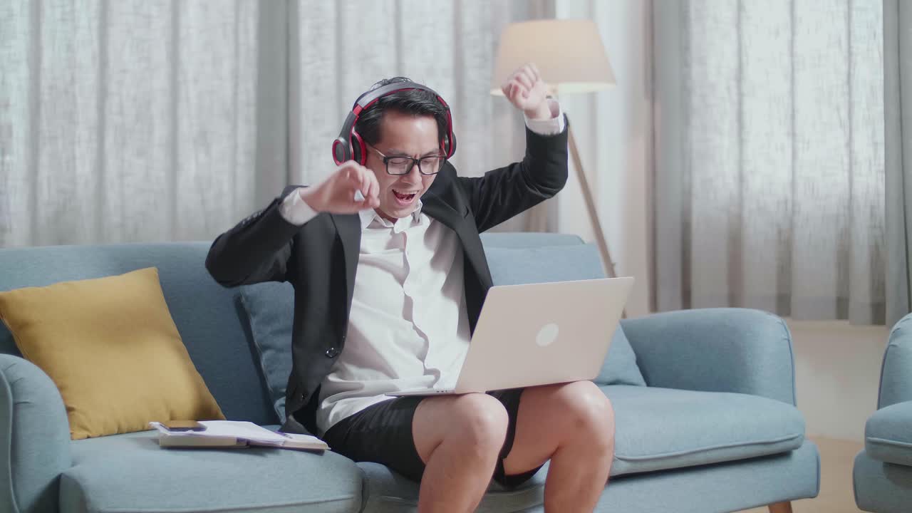 hombre de negocios asiático en chaqueta y pantalones cortos escuchando música con auriculares y bailando mientras trabaja con una computadora portátil en casa