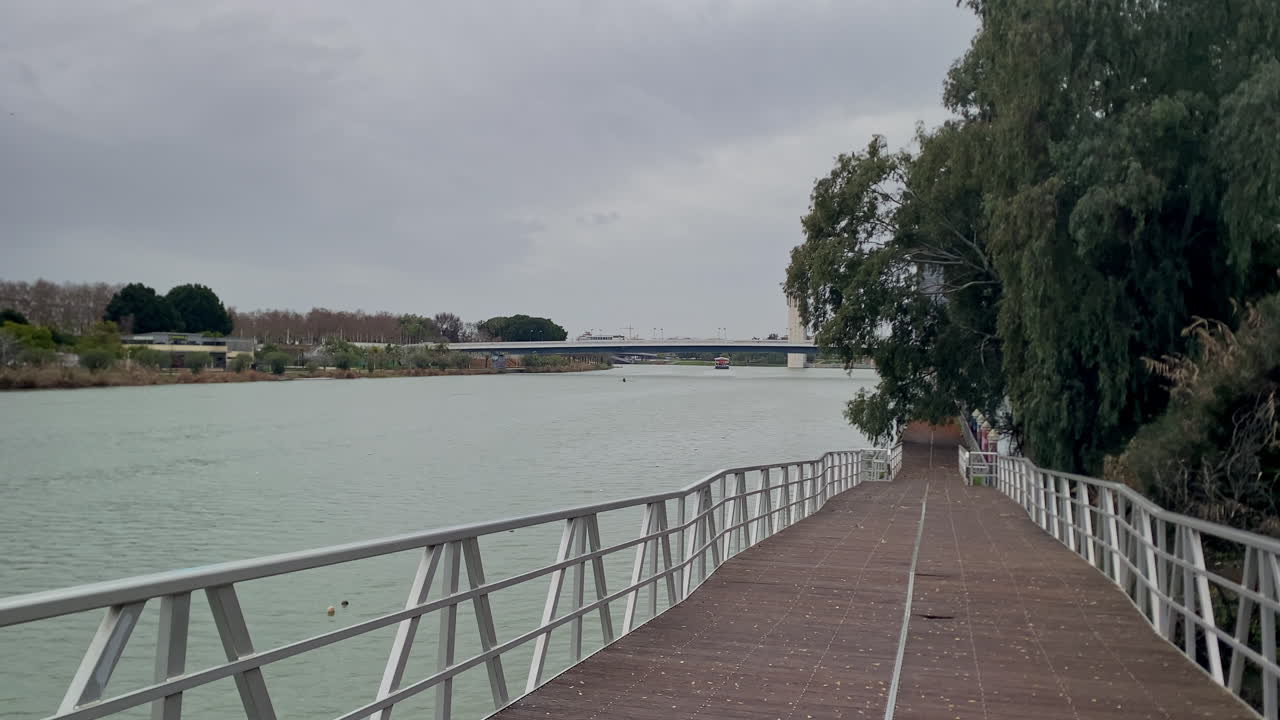 Řeka Guadalquivir protékající kolem moderní dřevěné nábřežní promenády a zelených stromů pod zataženou oblohou v Seville, Španělsko.