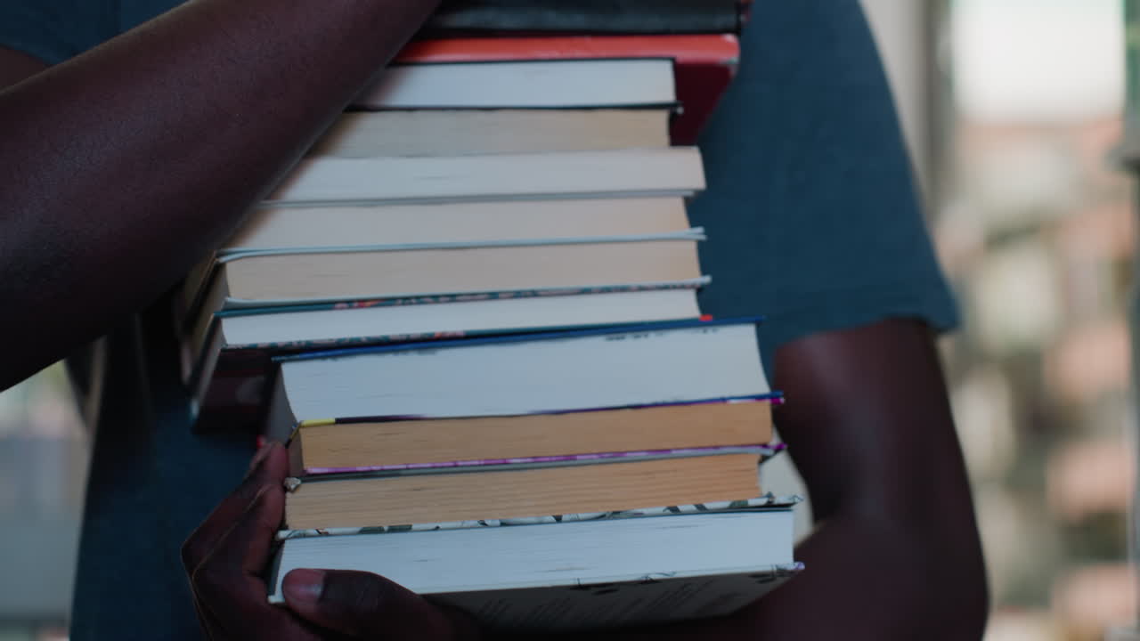 Close up of hands holding large stack of books while walking in library with blurred background, showing spines and pages in soft light, evoking academic dedication