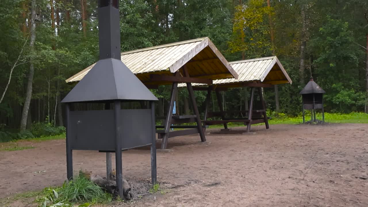Öffentlicher Waldgrillplatz mit überdachten Holzpicknickhütten und Metallgrills auf sandigem Boden, umgeben von Kiefern und Birken.