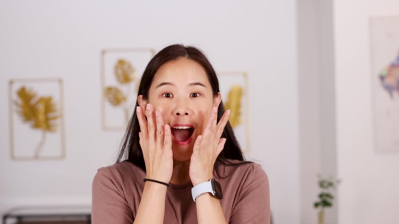 Asian woman expresses surprise and delight, hands on face, in bright, modern home interior