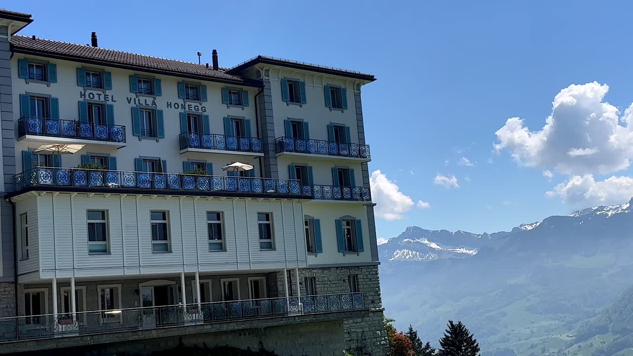 Hotel Villa Honegg in Switzerland with beautiful view on swiss alps and snowy peaks. Sunny summer day with green landscape and blue sky. Panorama panning wide shot. Luxury apartment resort.