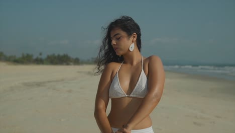 Südasiatische-Frau-Im-Weißen-Bikini,-Die-Am-Sonnigen-Strand-Sonnencreme-Auf-Die-Arme-Aufträgt