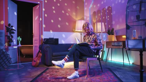 young stylish caucasian man with long hair and glasses sitting on a chair and reading a book in a retro room at home