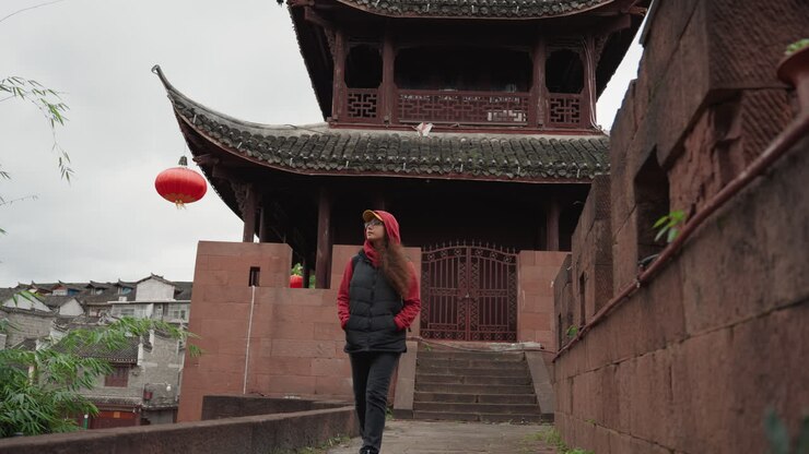 Femme asiatique montant les marches d'un temple sous des lanternes rouges, portant une doudoune noire et une casquette, ciel couvert, toile de fond de pagode aux tuiles brunes, murs et escaliers en pierre, humeur pensive, portrait de voyage et scène de rue