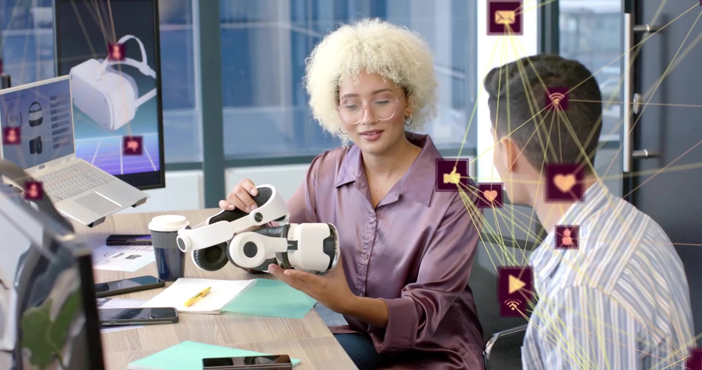 Woman tapping tablet showing animated network icons while team collaborating on VR headset designs