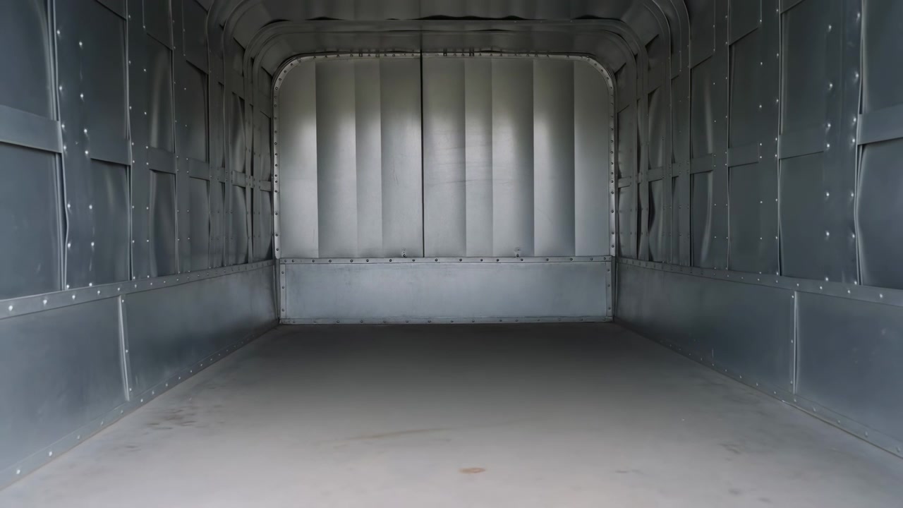 View pulls steadily away from vacant cargo area, exposing more of rear entrance while metallic side panels, curved roof framing, flat floor, and front wall remain centered and evenly lit.