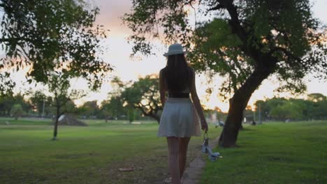 pretty young woman in hat walking barefoot carefree through park carrying shoes and rose in her hands