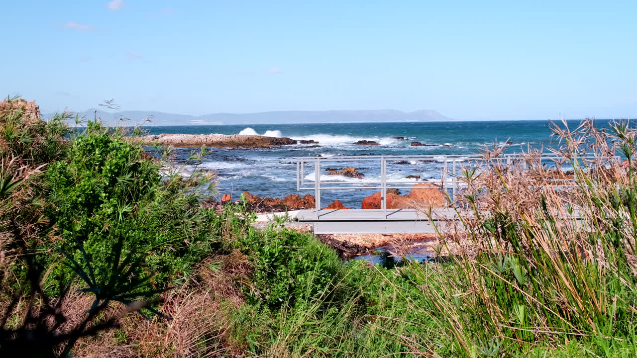 Man walks over small bridge on scenic coastal walkway on windy day, ocean behind