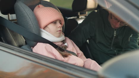 a man in a green jacket is carefully unbuckling the seat belt of his young daughter, who is calmly sitting in a car wearing a pink jacket and matching beanie