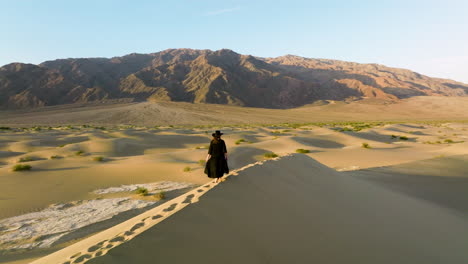 mujer con vestido negro y sombrero caminando sobre dunas de arena dorada dejando huellas